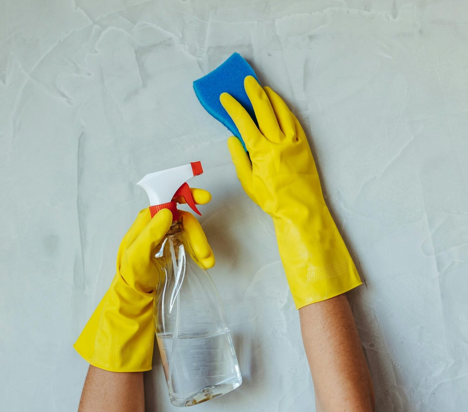 Close-up of hands in yellow gloves cleaning a wall with a spray bottle and sponge.