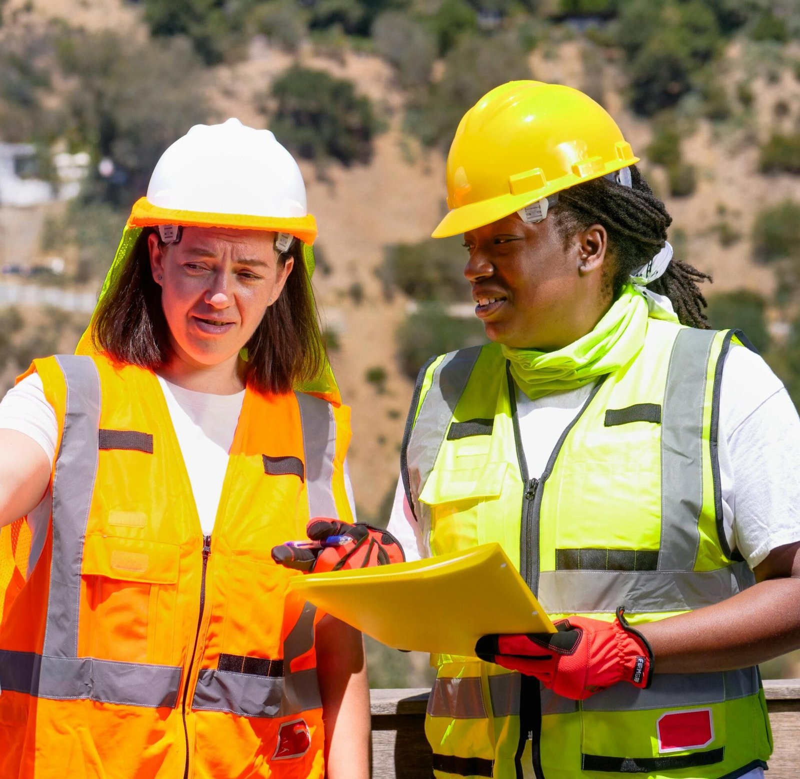 Two female engineers in safety gear discuss a project outdoors, focusing on plans.